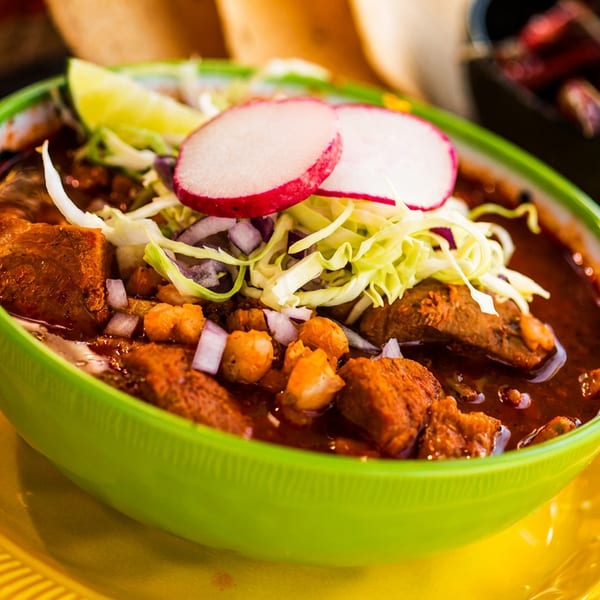 Vibrant green bowl of red pozole with meat, hominy, cabbage, radish, and lime.