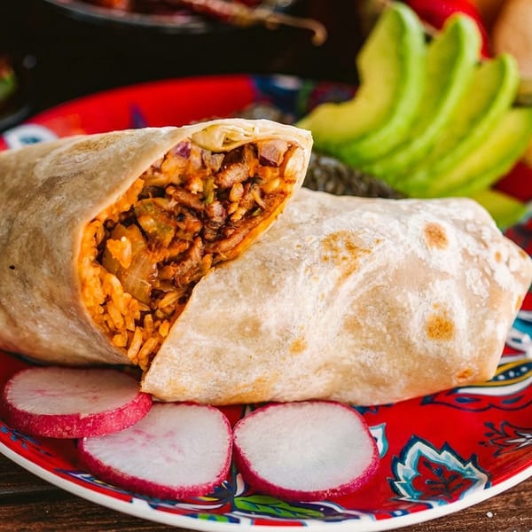 A large burrito, cut open, on a red patterned plate with radish slices and avocado.
