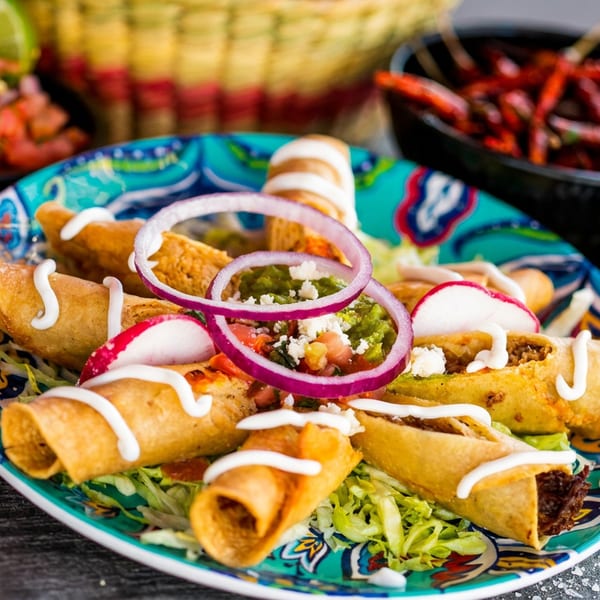 Colorful plate of crispy taquitos with lettuce, sour cream, onions, radishes, guacamole, and cheese.