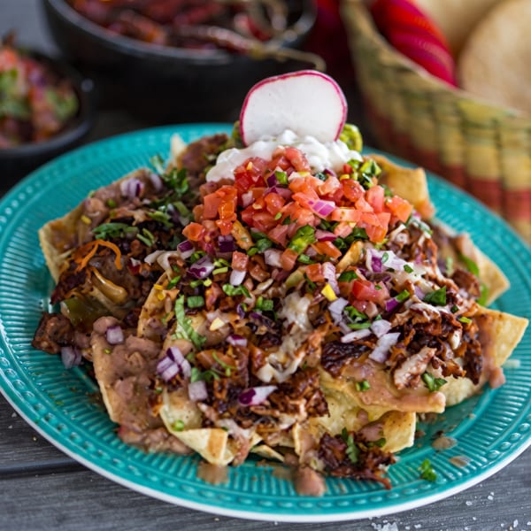 Teal plate of loaded nachos with pico de gallo, sour cream, radish, and meat.
