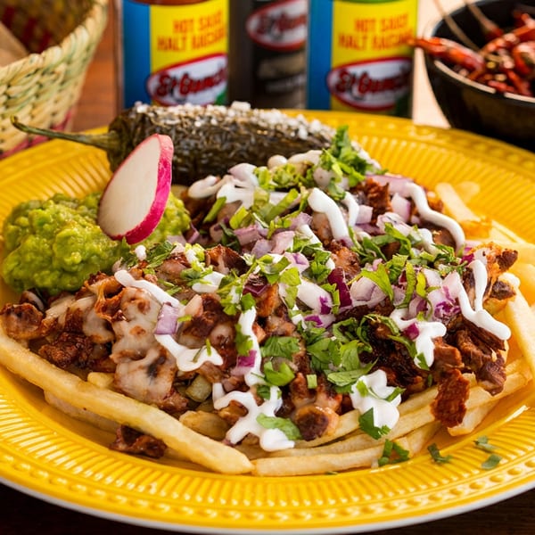 Loaded fries with meat, cheese, onions, cilantro, and crema on a yellow plate with guacamole and a jalapeño.